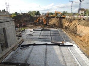 Geosynthetics trough under construction: Rolls of geogrid on top of drainage geocomposite in the foreground; geomembrane with anchoring grooves in the background.