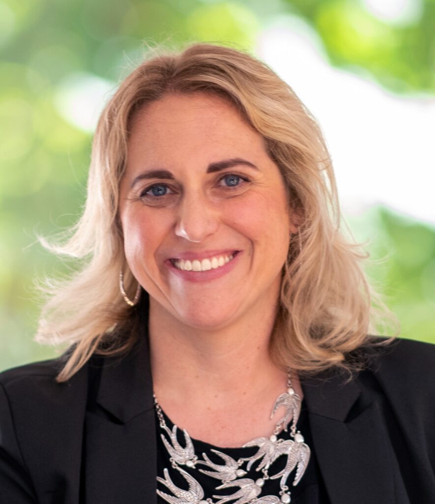 Smiling woman with long blonde hair, wearing a black blazer and a silver bird necklace, set against a green, blurred background.