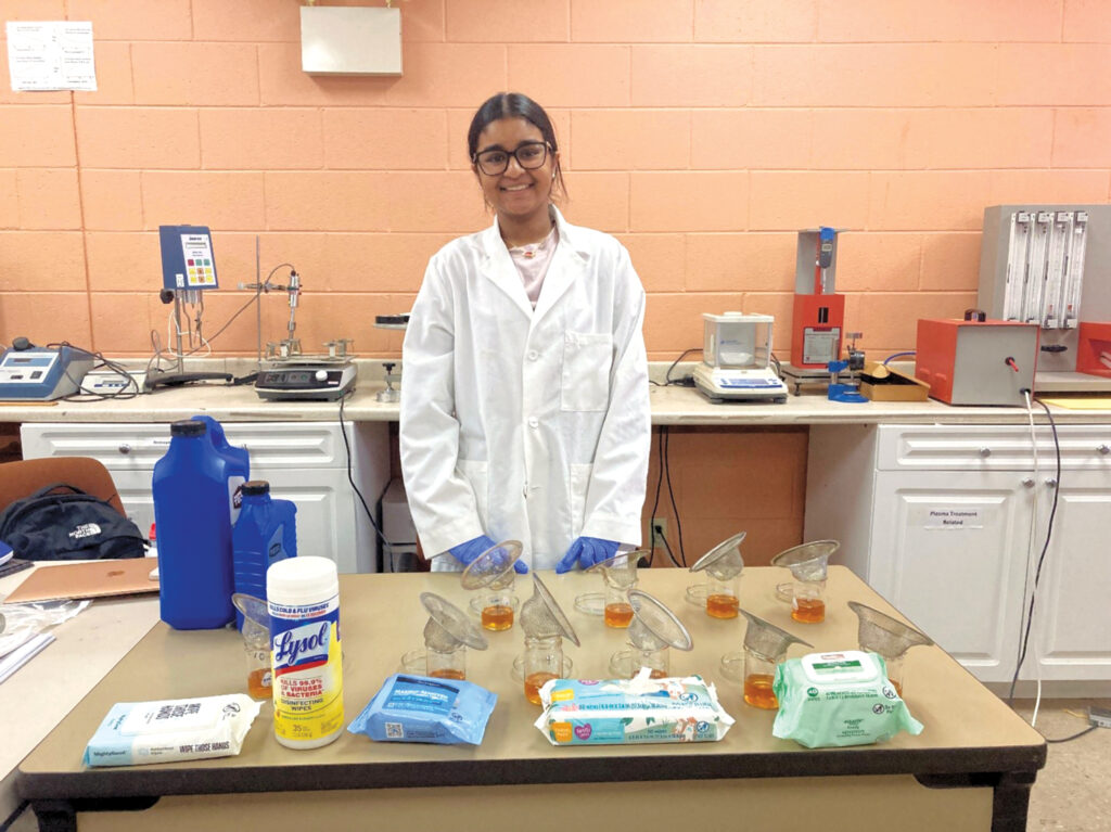 A young person in a lab coat stands confidently behind a table with beakers and various lab materials, in a bright classroom setting.