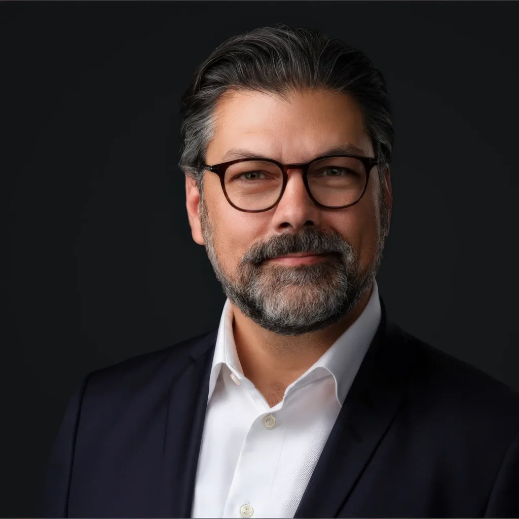 Man with a beard and glasses, wearing a dark suit and white shirt, smiles against a dark background.