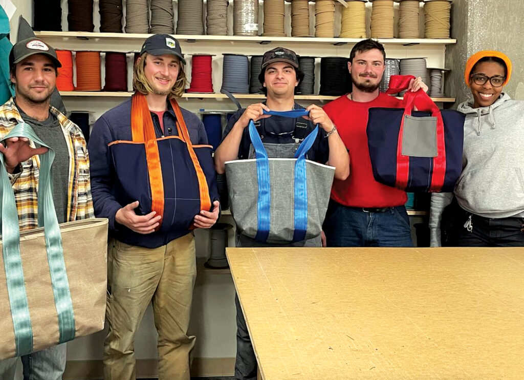 Five young adults pose with colorful tote bags in a workshop filled with spools of fabric. The atmosphere is vibrant and creative.