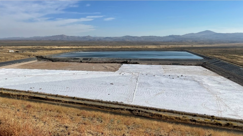 A vast landscape with a pond in the background, a white, textured area in the foreground, and mountains under a clear blue sky.