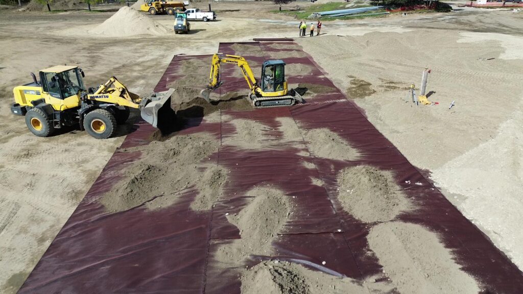 Construction site with yellow machinery on a maroon geotextile surface, featuring piles of sand and workers in safety vests nearby.