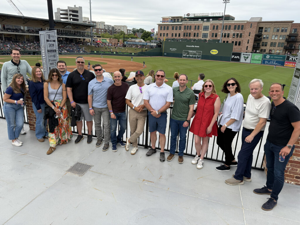 ATA Outlook Conference Group of professionals posing in front of a baseball field, with a mix of casual attire and vibrant backgrounds, enjoying an outdoor event.