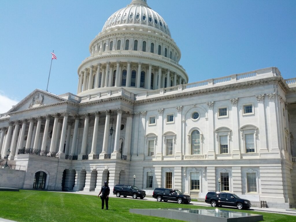 The U.S. Capitol building features a prominent dome and neoclassical architecture, with cars parked in front and a clear blue sky.