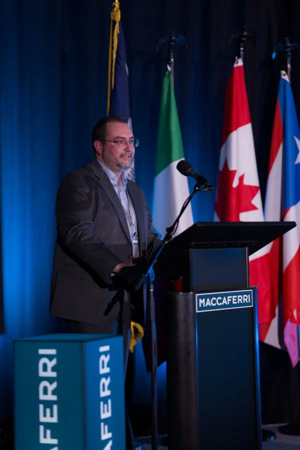 Man speaking at a podium, flanked by flags of Italy, Canada, and Texas, with a blue and black backdrop.