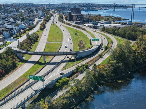 Aerial view of a highway with green trees along the sides, merging lanes, and a river in the background with buildings and a bridge.