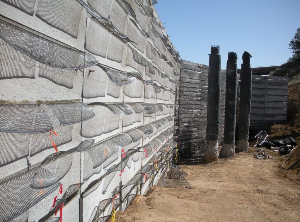 Geosynthetic-reinforced MSE wall under construction, featuring gray concrete elements, mesh layers, and vertical reinforcement columns.