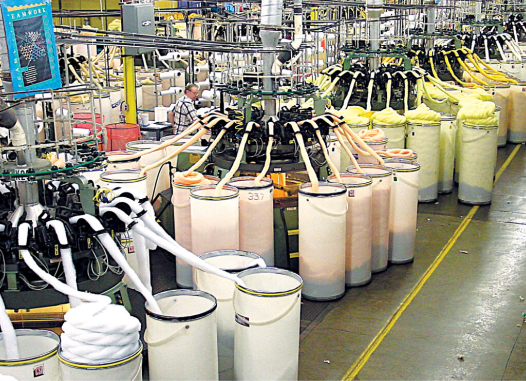 Industrial facility featuring rows of large, cylindrical containers with hoses, showcasing a production line with workers in the background.