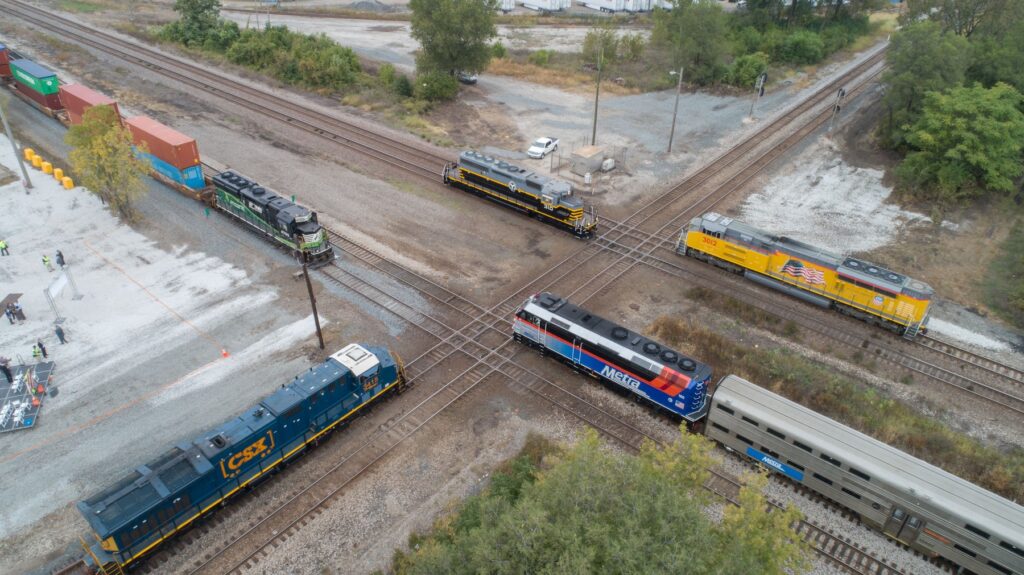 Aerial view of intersecting railway lines with multiple trains, including freight and passenger, surrounded by greenery and containers.