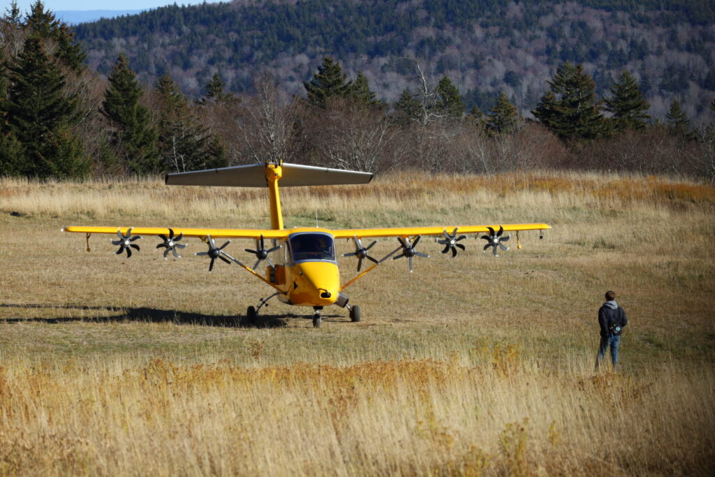 A bright yellow aircraft with multiple propellers on its wings is parked in a grassy field, while a person stands nearby, surrounded by trees.