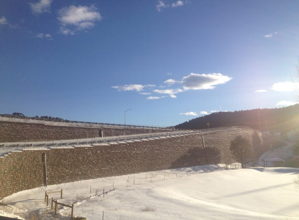 A snowy landscape with curved, terraced walls made of stone, against a blue sky with scattered clouds and a hint of sunlight.