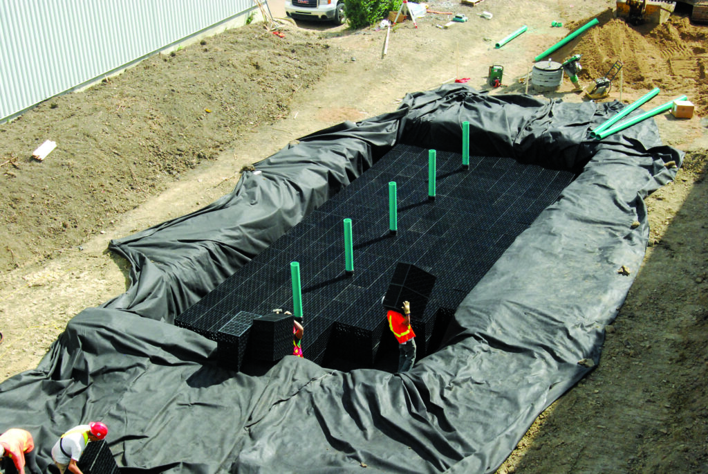 A large excavation site covered with black fabric, featuring green vertical pipes and workers arranging black modular drainage units.