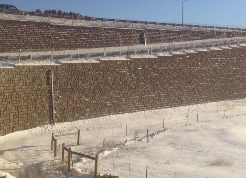 Geosynthetic-reinforced retaining wall in a snowy landscape, featuring layered stone blocks, with a highway visible above.