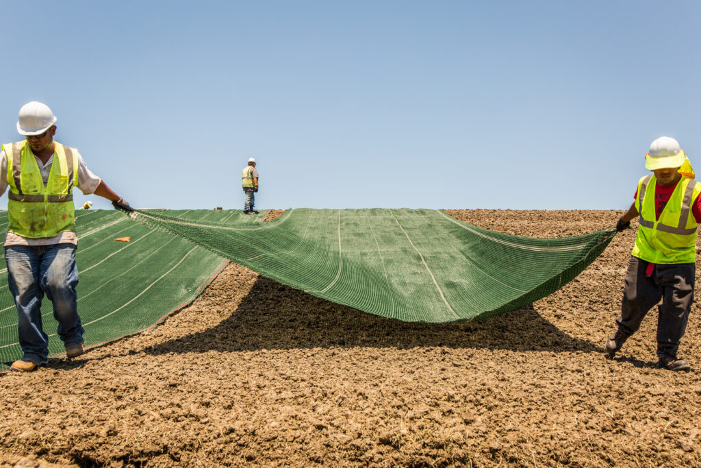 Two workers in hard hats and vests pull green erosion control fabric across a levee, with a third worker overlooking in the background.