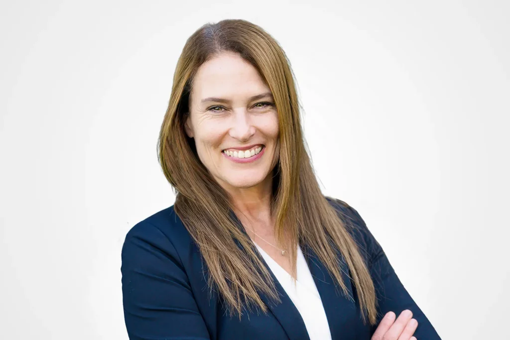 A smiling woman with long brown hair wearing a dark blazer and a white blouse, with arms crossed against a light background.