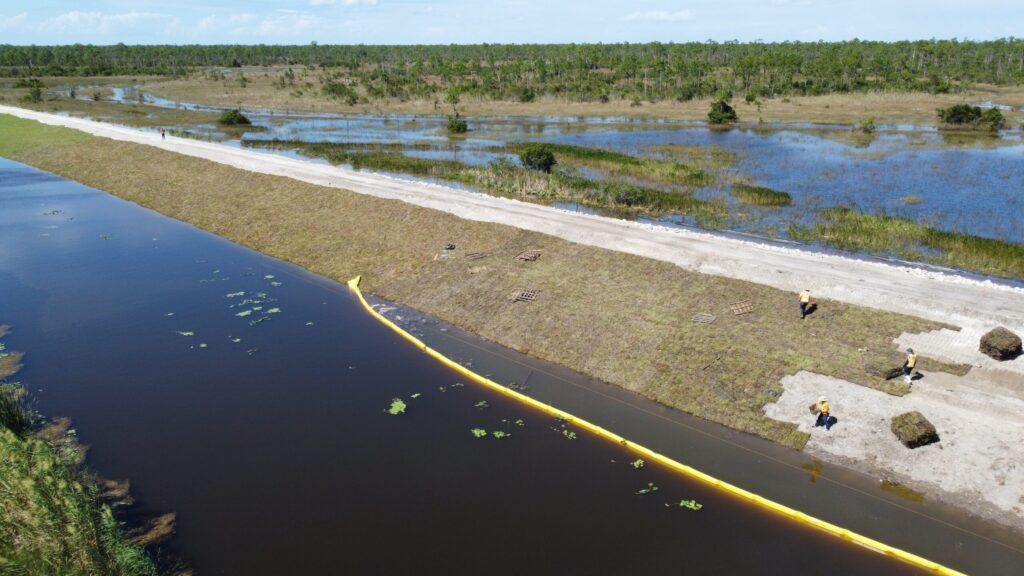 Aerial view of a levee under construction with workers, a muddy waterway, and grassy banks, surrounded by wetlands and trees.