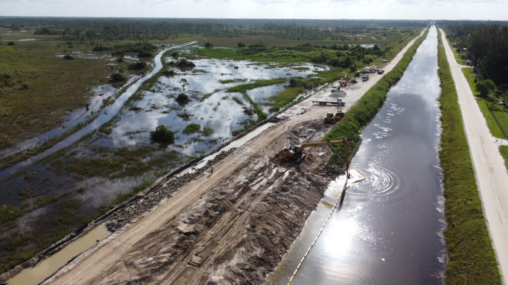Aerial view of a levee construction site alongside a waterway, featuring heavy machinery and vegetated marshland under a clear sky.
