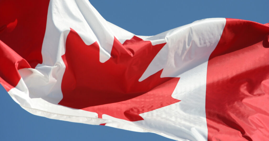 Canadian flag fluttering against a clear blue sky, prominently displaying the red and white colors with a central maple leaf emblem.