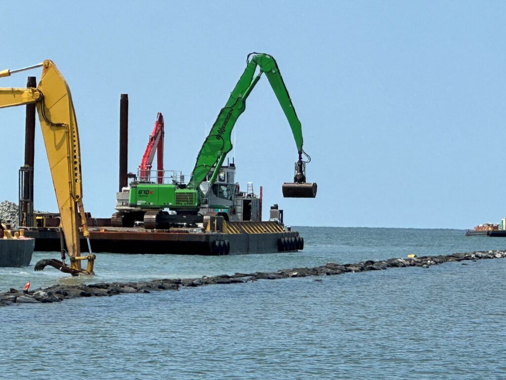 A green excavator lifts a large object over a barge, with yellow and red machinery in the background against a clear blue sky.