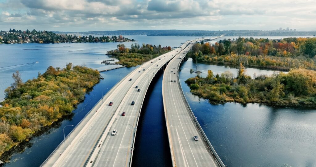 Aerial view of a divided highway crossing water, surrounded by colorful trees and greenery under a partly cloudy sky.