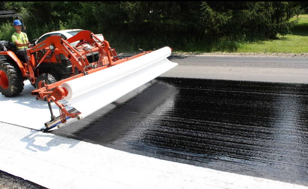 A tractor applies a black liquid material onto a roadway, with a wide white roller in the foreground and greenery in the background.