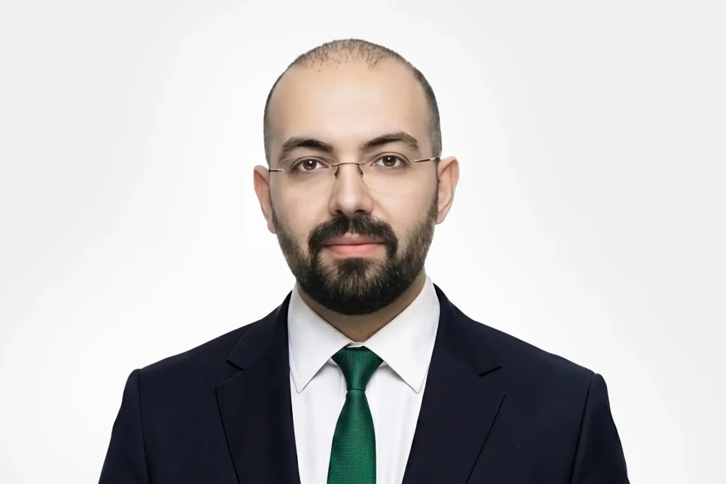 Man with a beard and glasses, wearing a dark suit, white shirt, and green tie, against a light gradient backdrop.