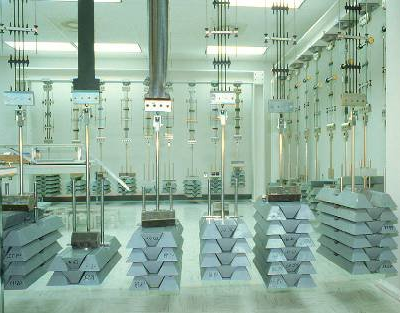 Laboratory setup featuring metal weights stacked on rectangular bases, suspended by cables, with testing equipment in a bright, organized space.