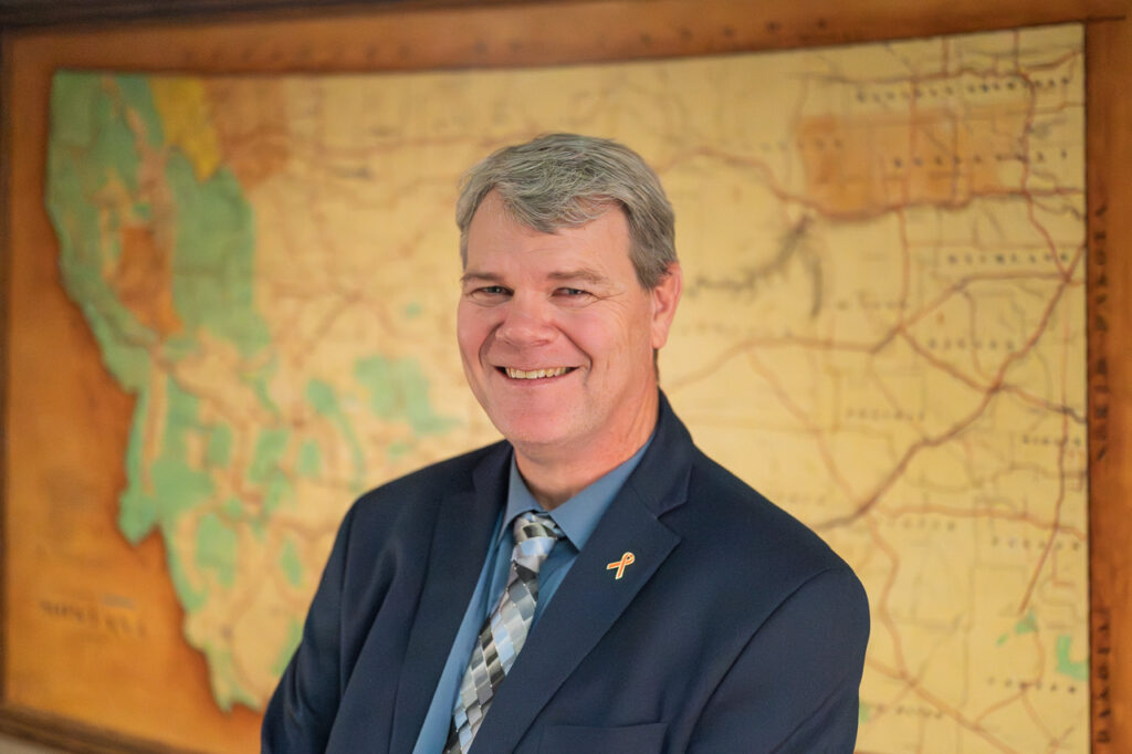 Smiling man in a dark suit with a patterned tie stands in front of an antique map, featuring shades of brown and green.