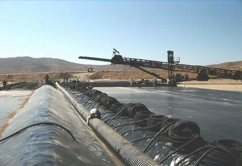 Black geomembranes cover a mining site, with a conveyor system in the background and workers present under a clear sky.
